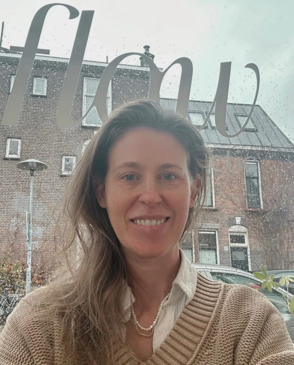 Een vrouw met lang, golvend bruin haar glimlachend voor een raam met de tekst 'fLnt' erop, met een gebouw en een straat te zien op een regenachtige dag.