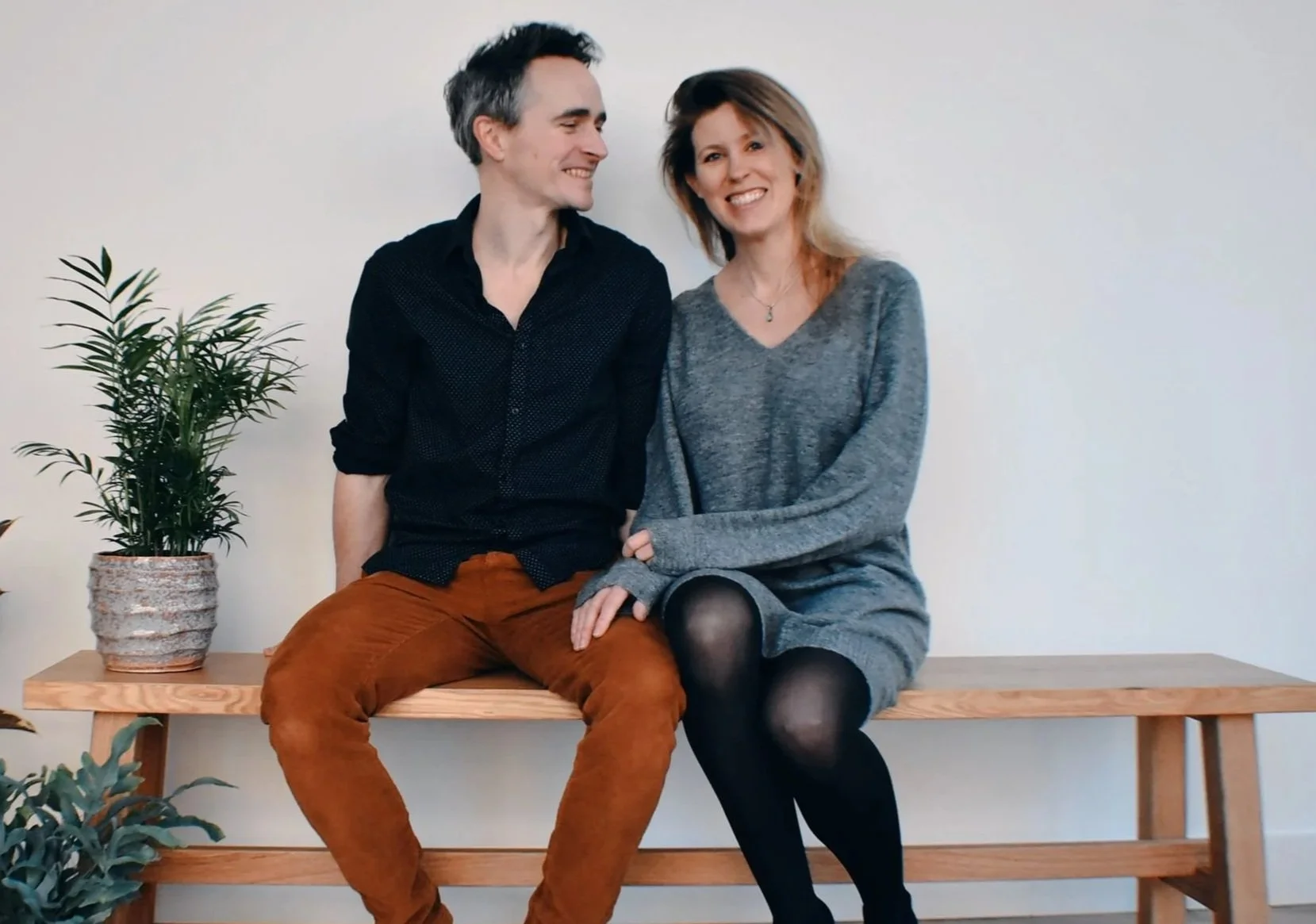 Een man en een vrouw zitten samen op een houten bank met planten op de tafel ernaast, glimlachen en kijken elkaar aan in een lichte kamer.