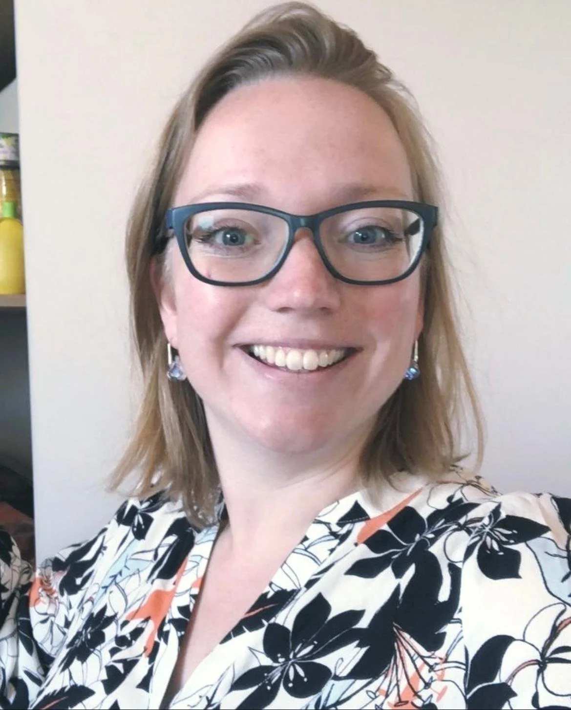 Een glimlachende vrouw met bril en oorbellen, draagt een zwart-wit bloemenpatroon shirt, tegen een witte muur.