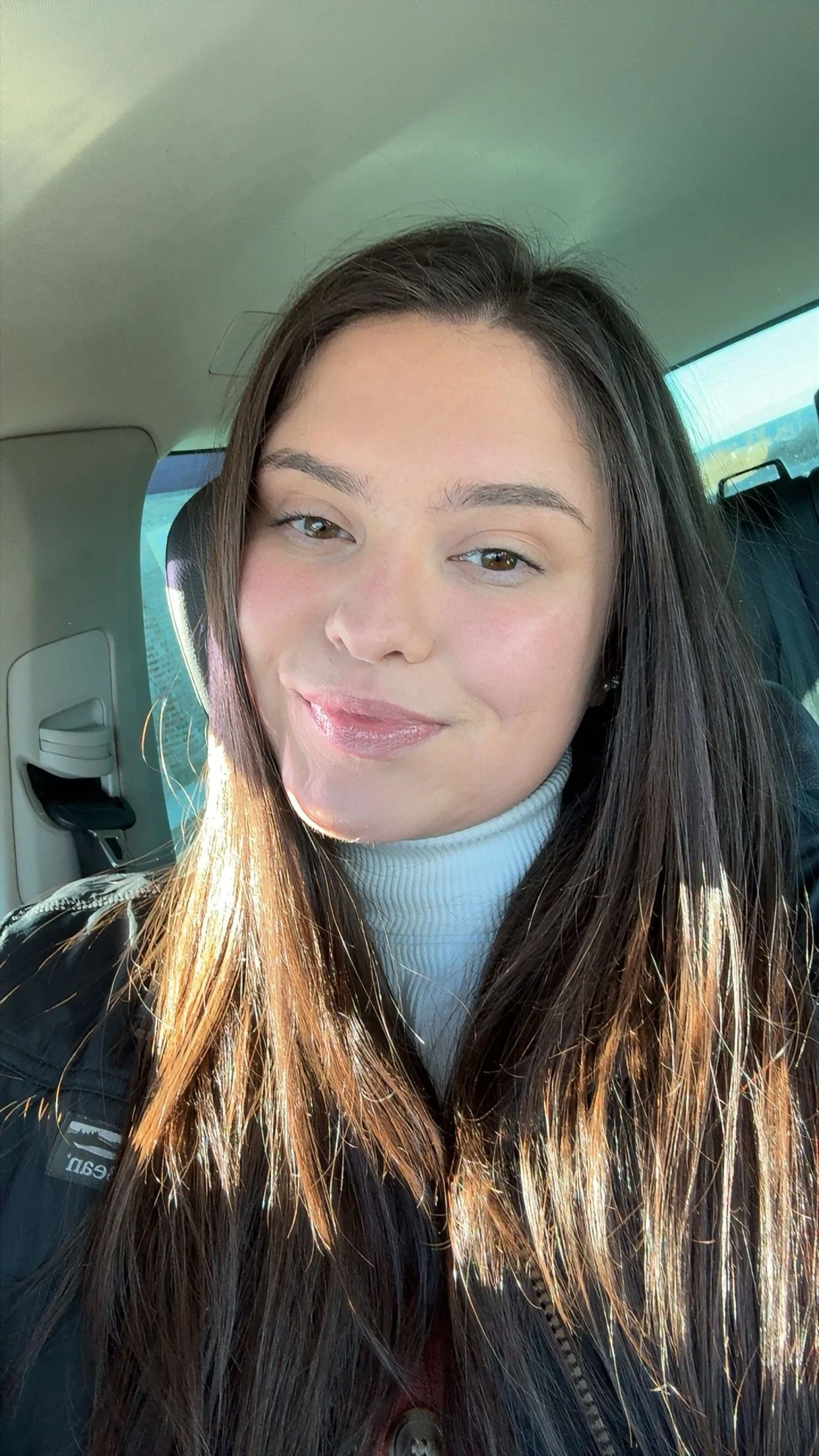 A young woman with long dark hair smiling, wearing a navy blue polo shirt with embroidered text and a small gold necklace, against a gray background.
