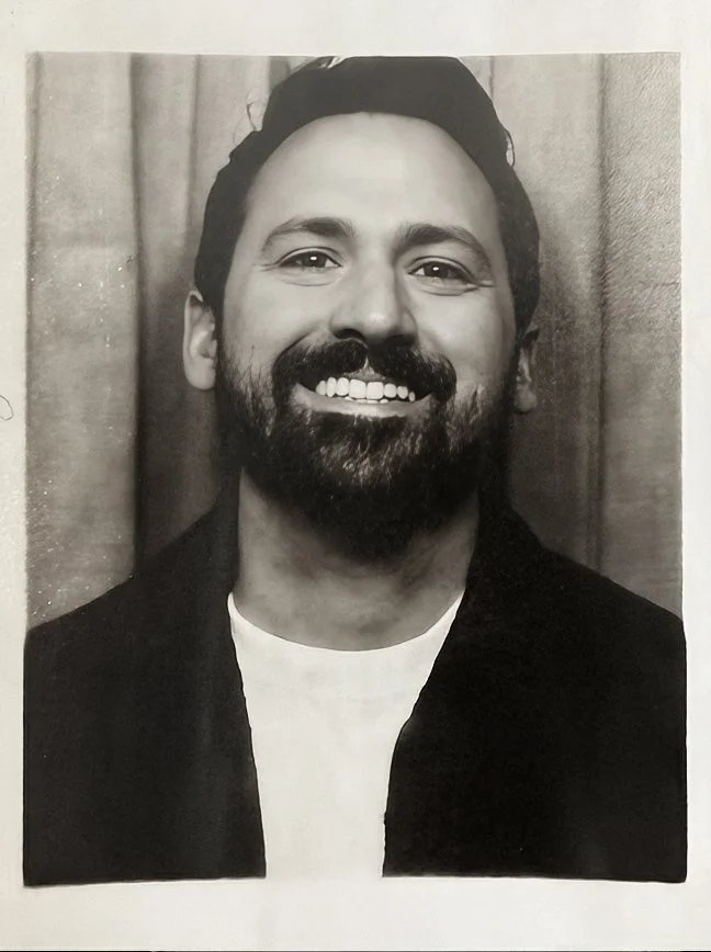 A smiling man with dark hair, a beard, and a mustache standing outdoors near a body of water.