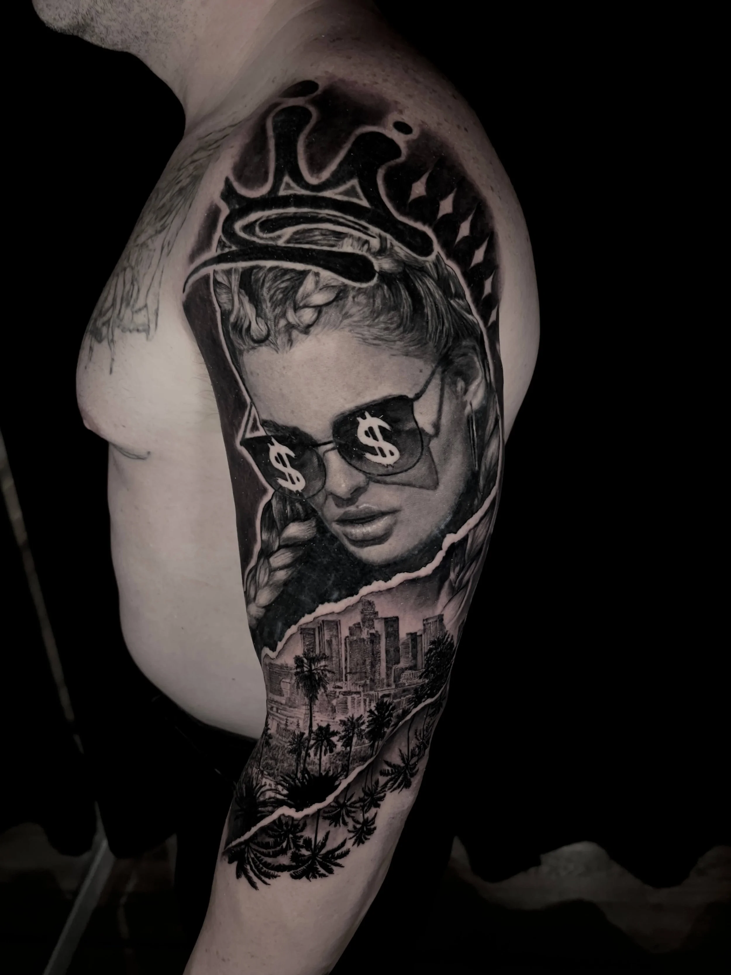 Woman in dollar-sign sunglasses with crown and city skyline, realism tattoo on the upper arm by Etgar Oak