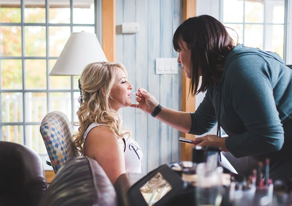 Makeup artist applying lipstick to a bride sitting in a chair with natural light coming through windows.