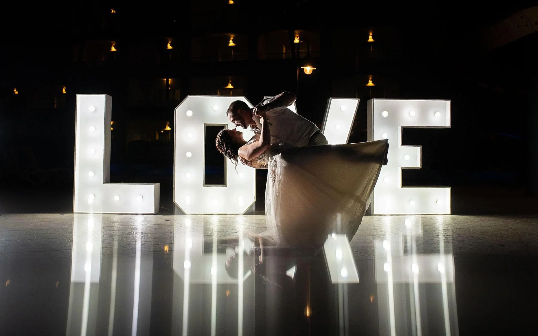 A bride and groom dancing in front of large illuminated letters spelling "LOVE" at a wedding reception, with their reflection visible on the shiny floor.