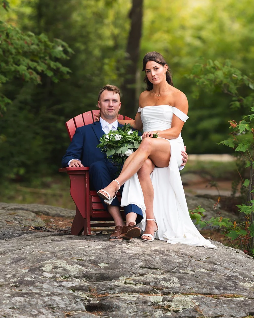 Bride and groom sharing a red Muskoka chair on the rocks at Armishaw Lake
