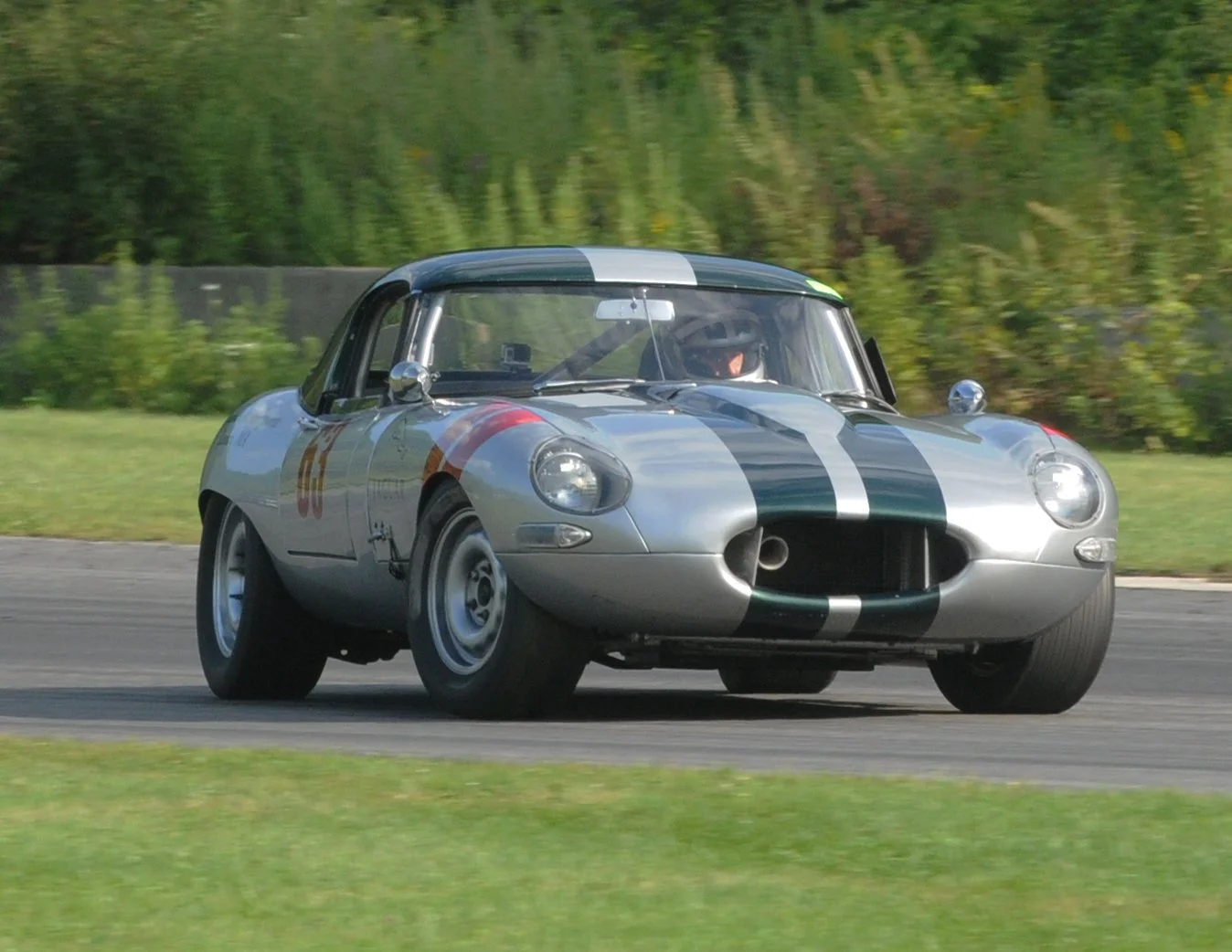 A classic silver racing car with black and green racing stripes on a race track, moving at high speed.