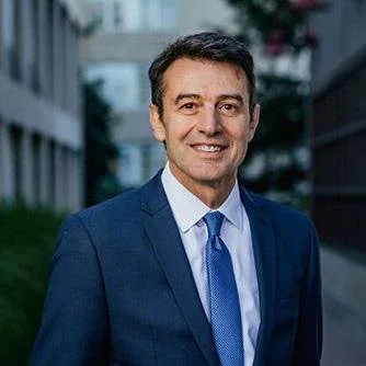 A man in a blue suit and tie smiling outdoors with buildings and trees in the background.