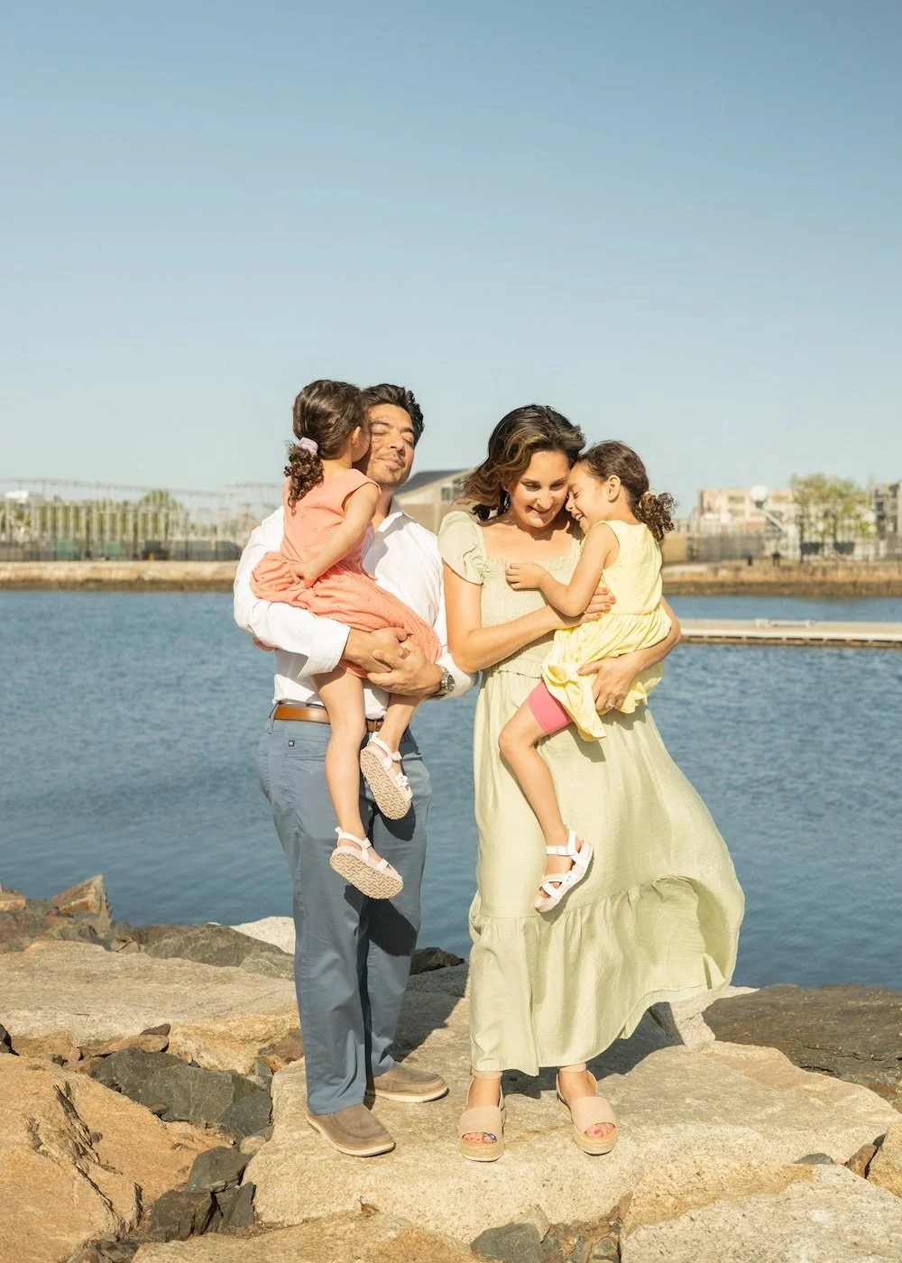A family of four standing on rocks by the water during daytime. The father is holding a young girl, and the mother is holding another girl. Both girls are hugging and kissing their parents, and everyone is smiling, enjoying a sunny day by the water.