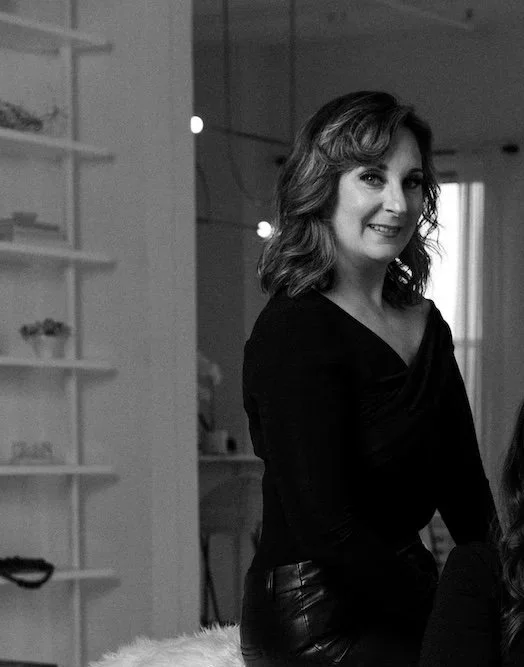 A woman with wavy shoulder-length hair smiling in a black top and leather pants, indoors with shelves and curtains visible in the background.