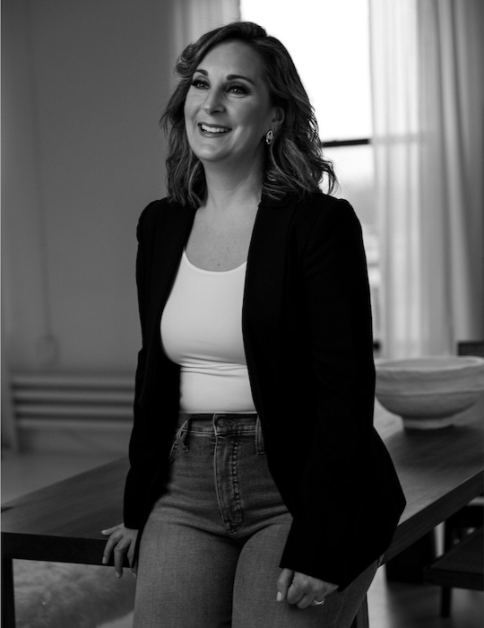 A smiling woman with shoulder-length wavy hair, wearing a black blazer over a white top, standing in a room with large windows and a wooden table.
