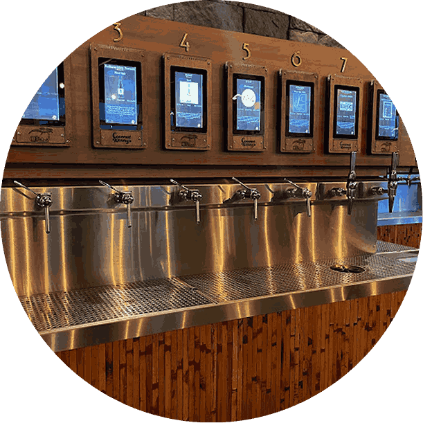 A self-serve beverage station with seven touch-screen screens labeled 3 to 7 above metal dispensers, in a restaurant or cafe setting.