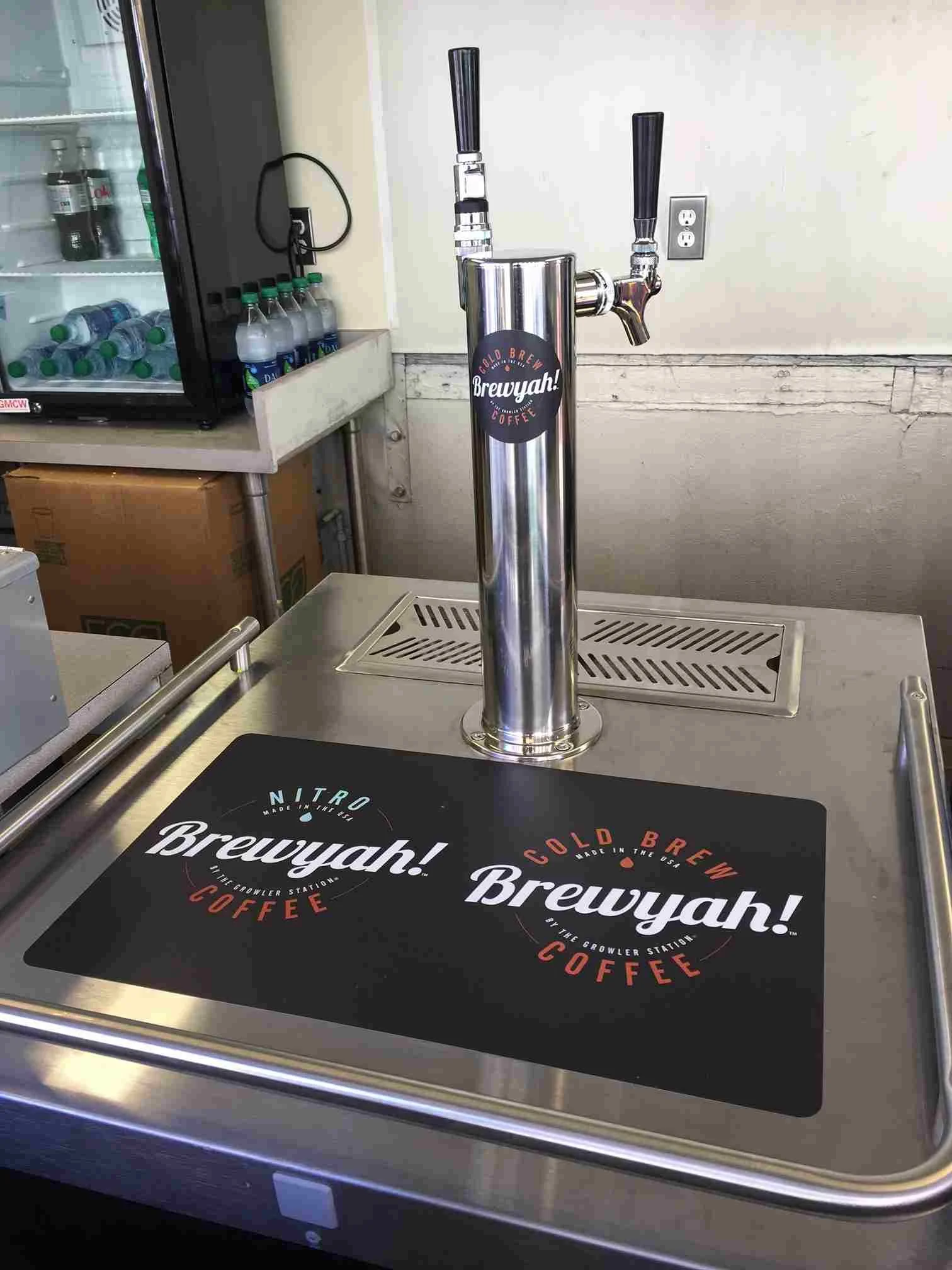 Stainless steel draft beer tap with two handles, labeled "Brewyah!" and "Cold Brew," on a countertop with a branded mat. A refrigerator with bottled drinks is in the background.
