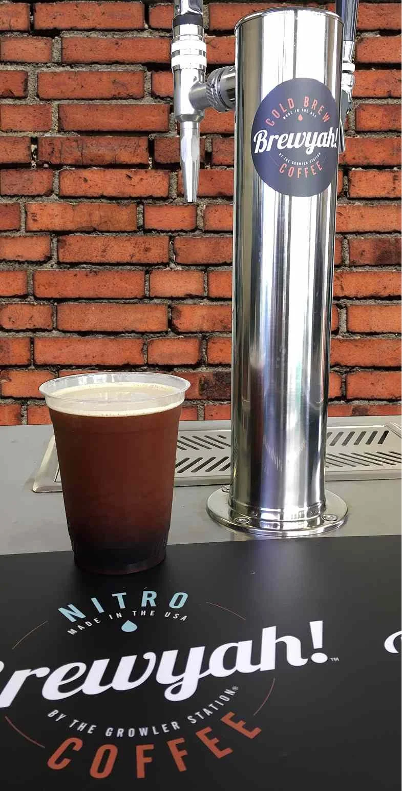 A coffee cup filled with cold brew coffee in front of a stainless steel cold brew coffee tap system against a brick wall.