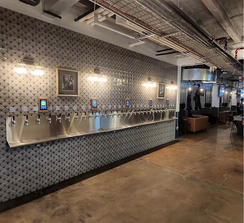 A self-serve tap wall lined up along a textured gray tiled wall and framed pictures, in a modern bar or brewery.
