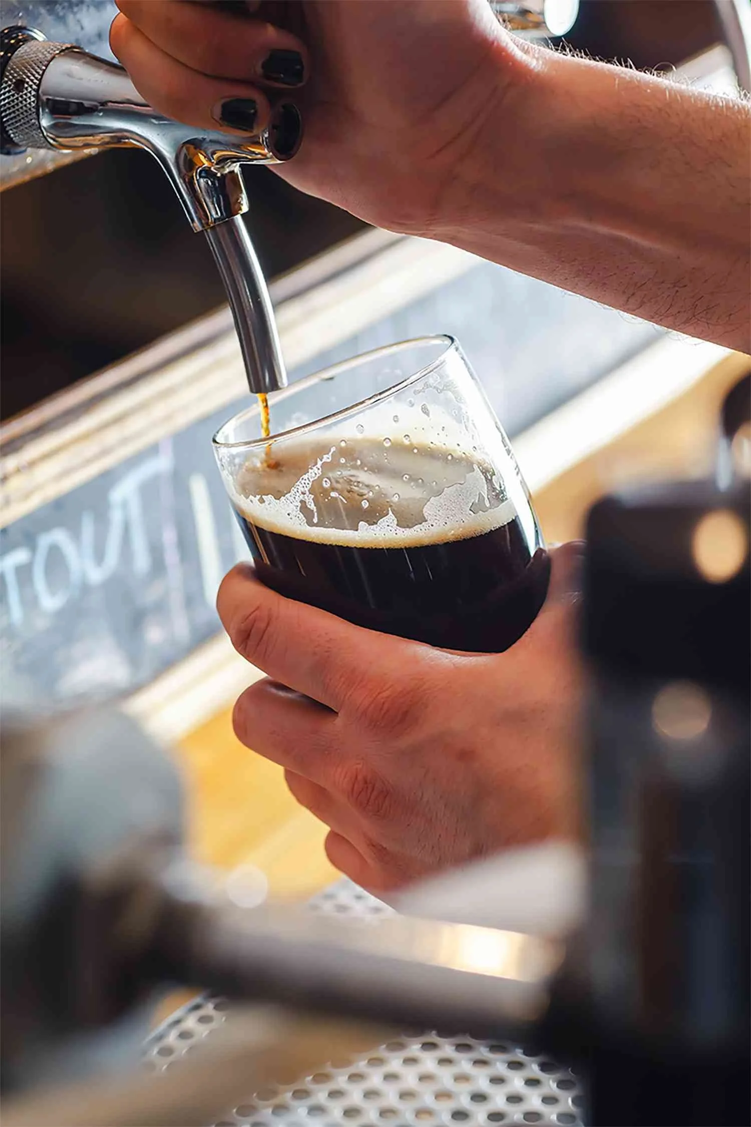 A hand holding a glass below a tap pouring dark beer. The tap is attached to a metallic dispenser, with a blurred chalkboard in the background.