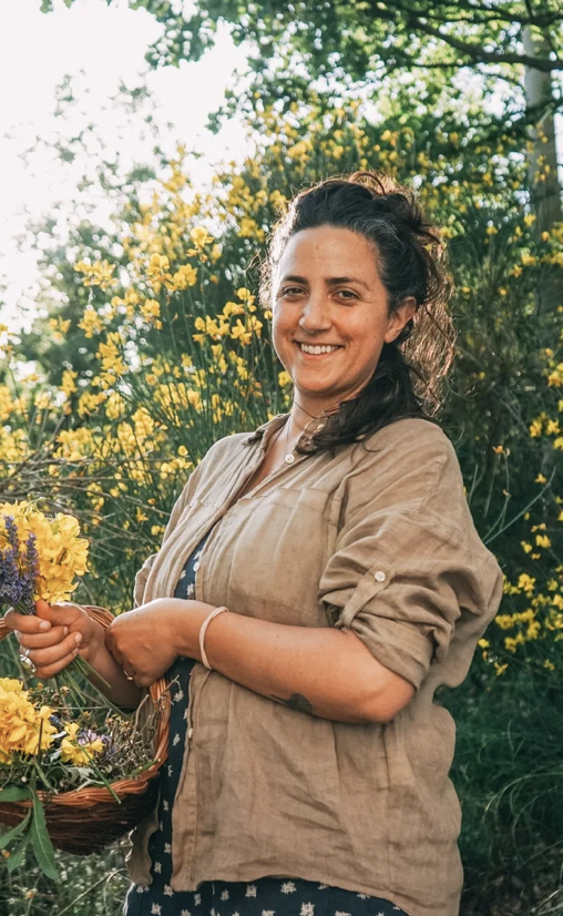 Una donna sorridente che tiene un cesto di fiori con sfondo di alberi e fiori gialli, in un ambiente all'aperto soleggiato.