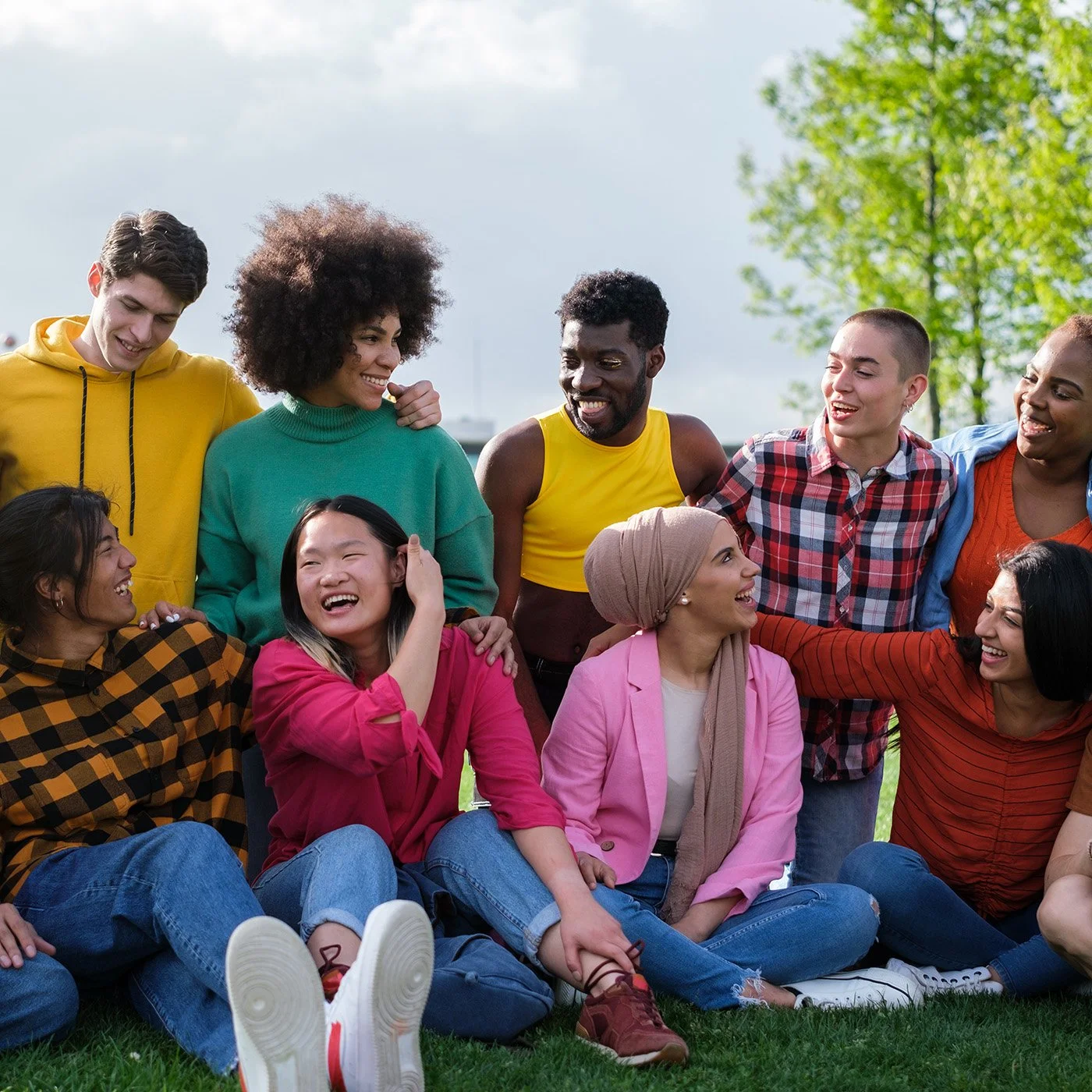 A diverse group of people joyfully smiling and talking outdoors on a sunny day, sitting and standing on grass with trees in the background.