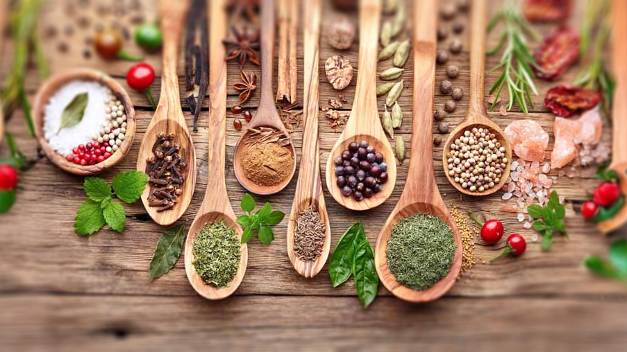 Assorted spices in wooden spoons on a wooden surface, including salt, peppercorns, turmeric, coriander, bay leaves, and herbs, with some cherry tomatoes, Himalayan salt, and fresh herbs around.