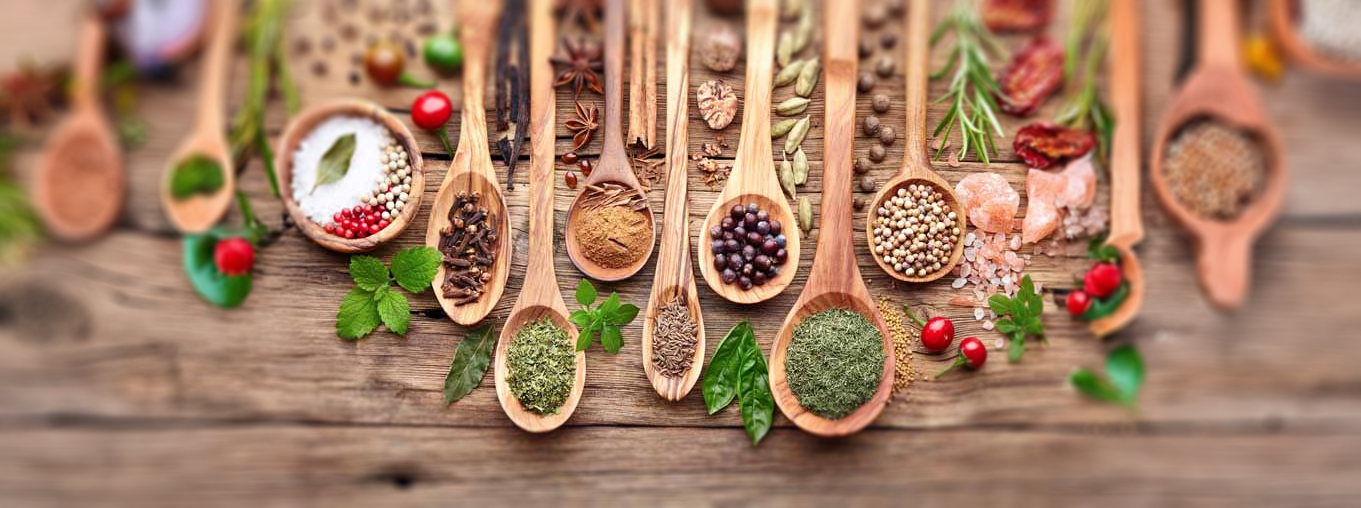 Various herbs, spices, and seasonings arranged on wooden spoons and scattered on a rustic wooden surface, including salt, pepper, cinnamon, cloves, and dried herbs.