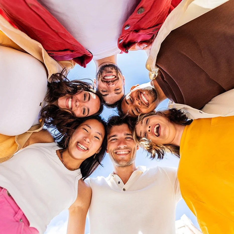 Group of six friends smiling and looking down at the camera, forming a circle outdoors with a blue sky background.