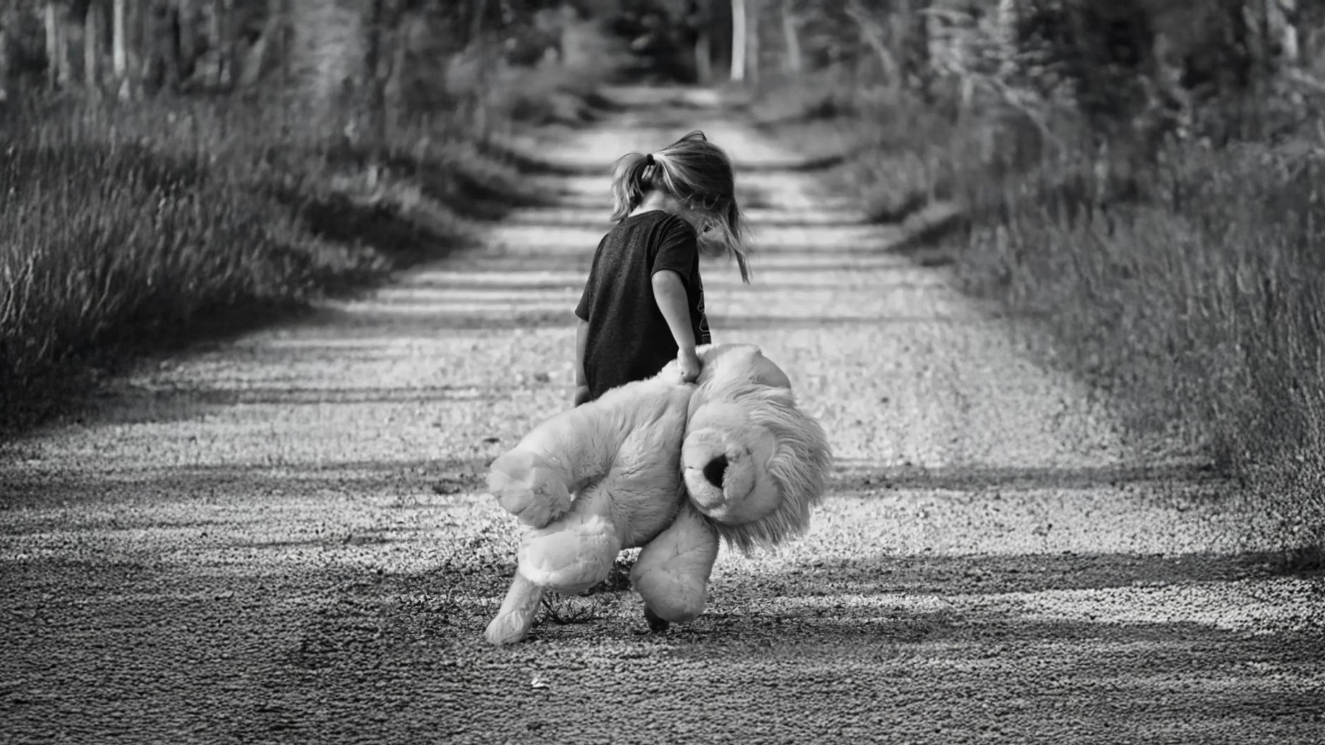 A young child walking down a dirt road holding a large stuffed animal, a plush dog, on their left side, on a rural path flanked by trees and grass, black and white photo.