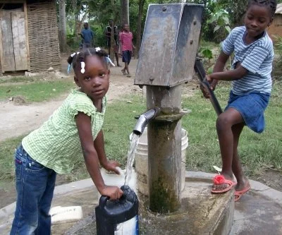 Two children collecting water from a community hand pump in a rural area, with one girl filling a container and a boy operating the pump.