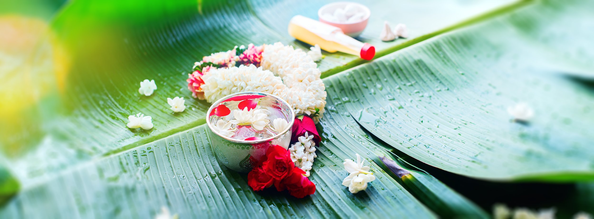 A traditional floral arrangement with white, pink, and red flowers on large green banana leaves, including a small bowl of water with floating flower petals, surrounded by flower petals and a tube of white cream or ointment.