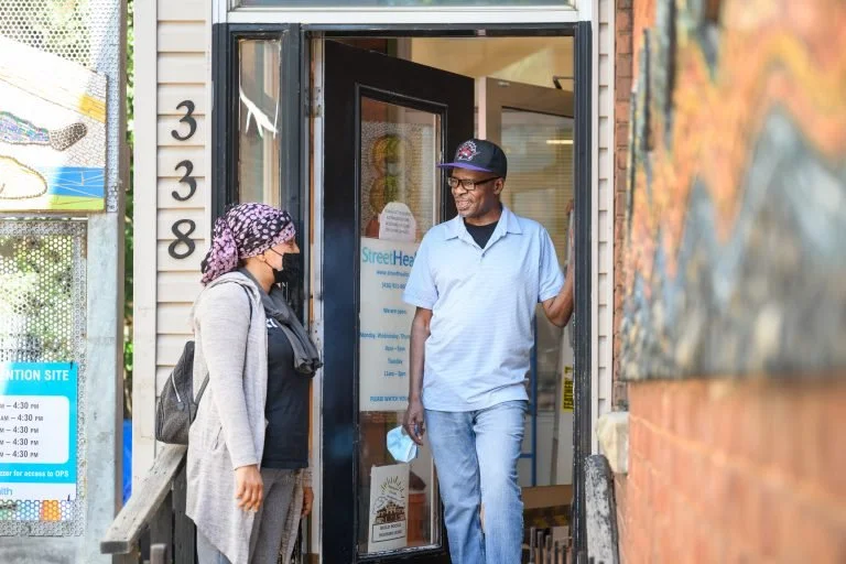 A man and woman are chatting at the doorway of a building, which appears to be a community health center. The man is standing in the doorway, smiling, wearing a baseball cap, glasses, a short-sleeve button-up shirt, and jeans. The woman, wearing a face mask, a headscarf, and a backpack, is standing outside the entrance.