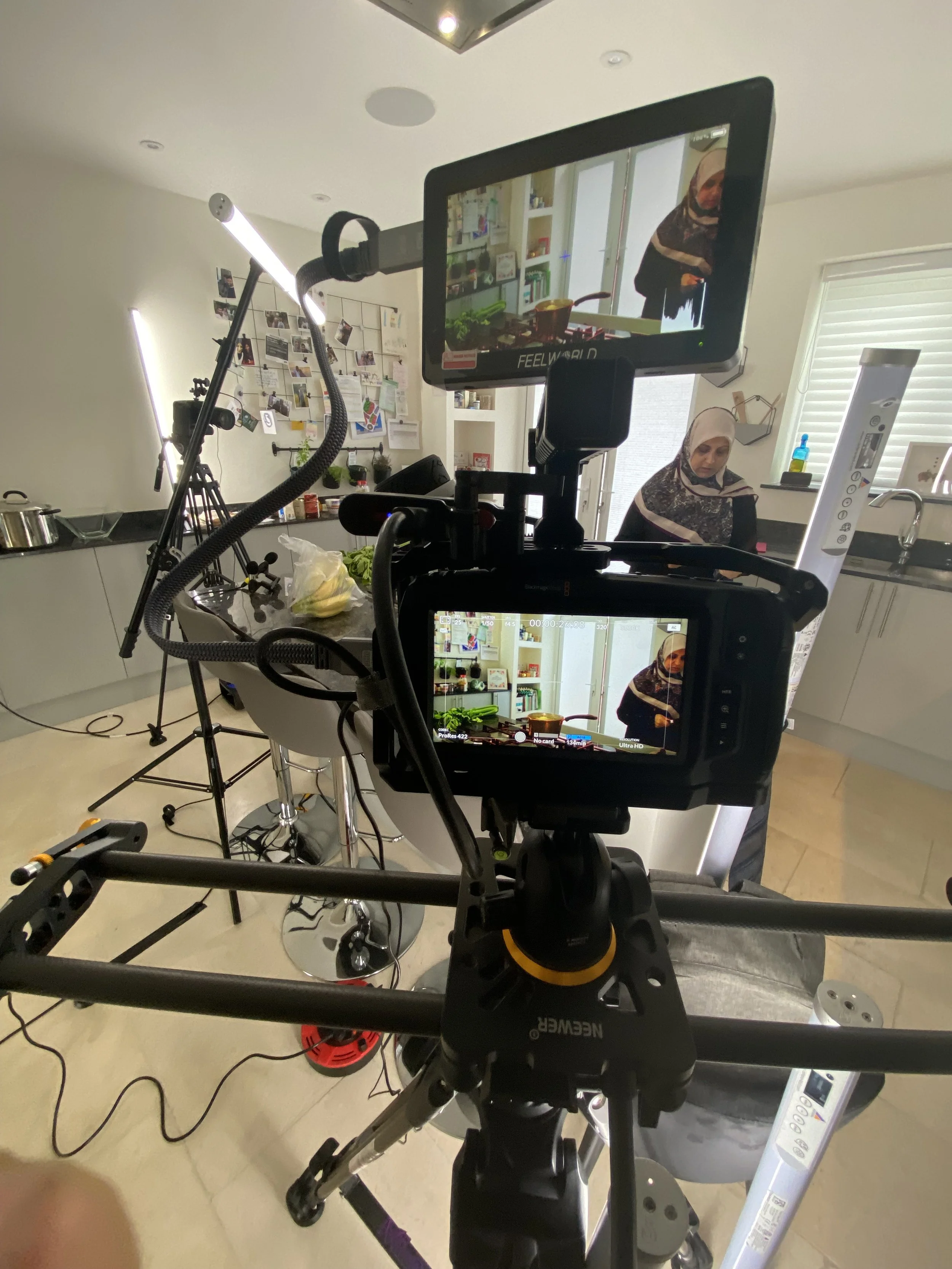 A woman in a kitchen, appearing on a video recording or live stream, surrounded by cameras and studio equipment, with a white cabinet, a black countertop, and kitchen items including a pot, fruit, and various containers in the background.