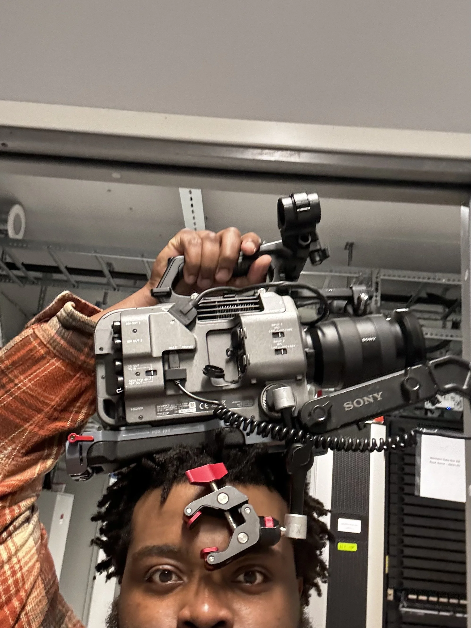 A person with dreadlocks is holding a professional video camera on their shoulder, in an indoor setting with electronic equipment in the background.