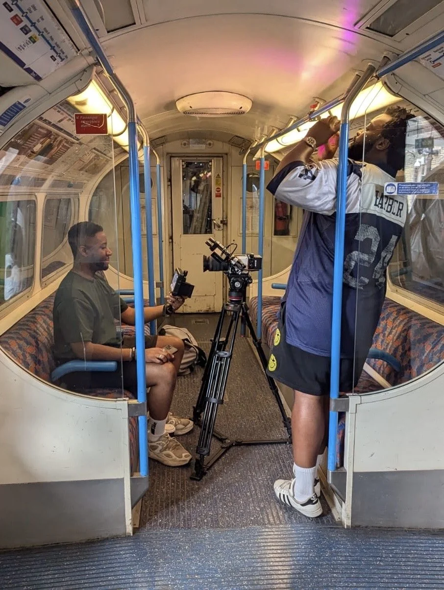 Two men filming an interview inside a vintage-style train carriage. One man is sitting on the left, holding a small camera, and the other is standing on the right, adjusting his shirt near a professional camera mounted on a tripod.