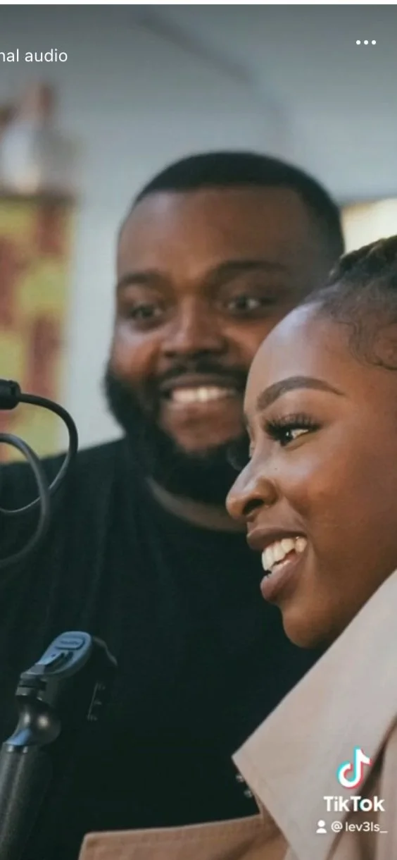 A man and a woman are smiling and looking at a microphone. The man has a beard and short hair, and the woman has styled hair and earrings.