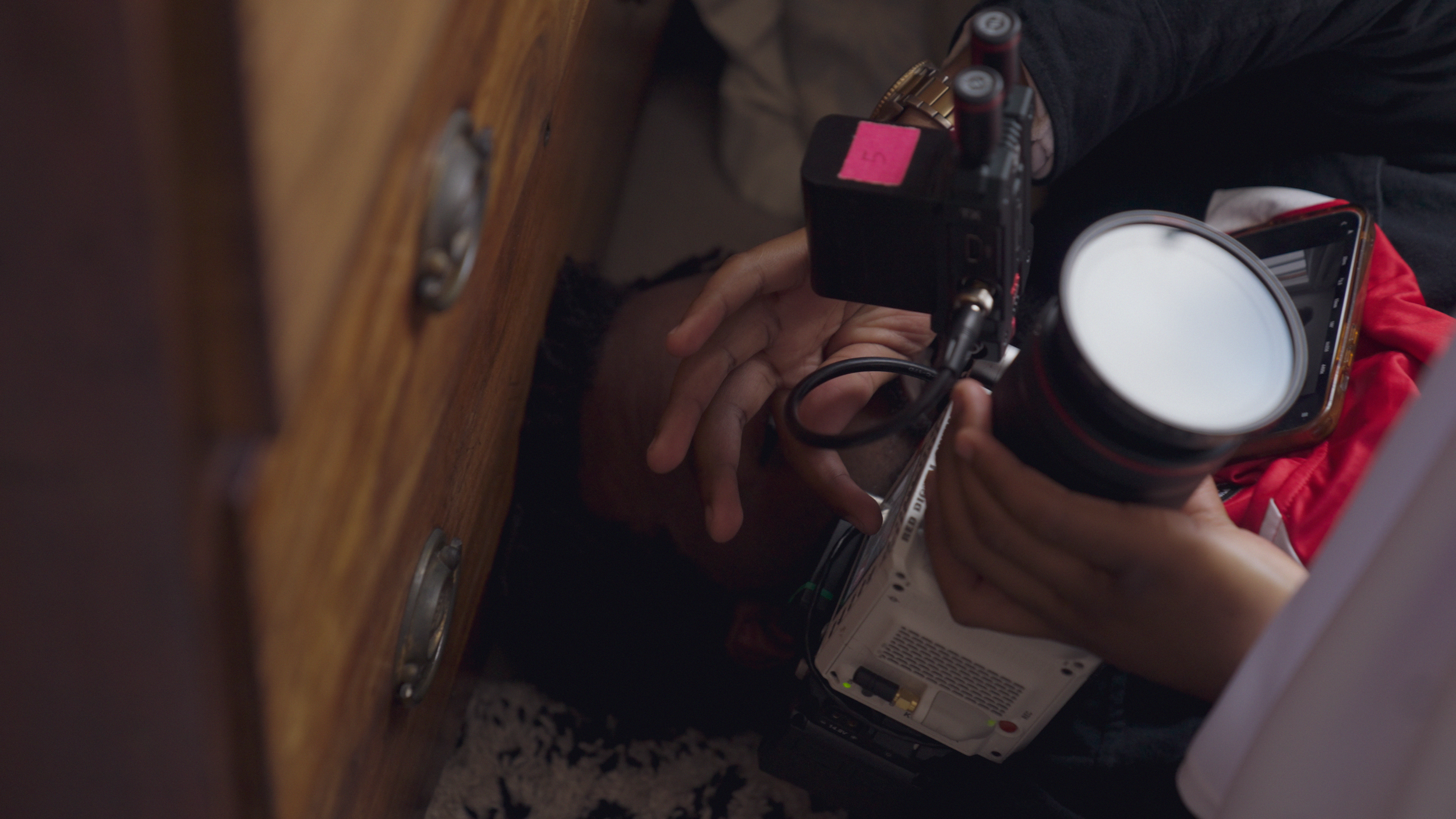 A person holding a camera, smartphone, and a white cylindrical object, with a wooden piece of furniture nearby.