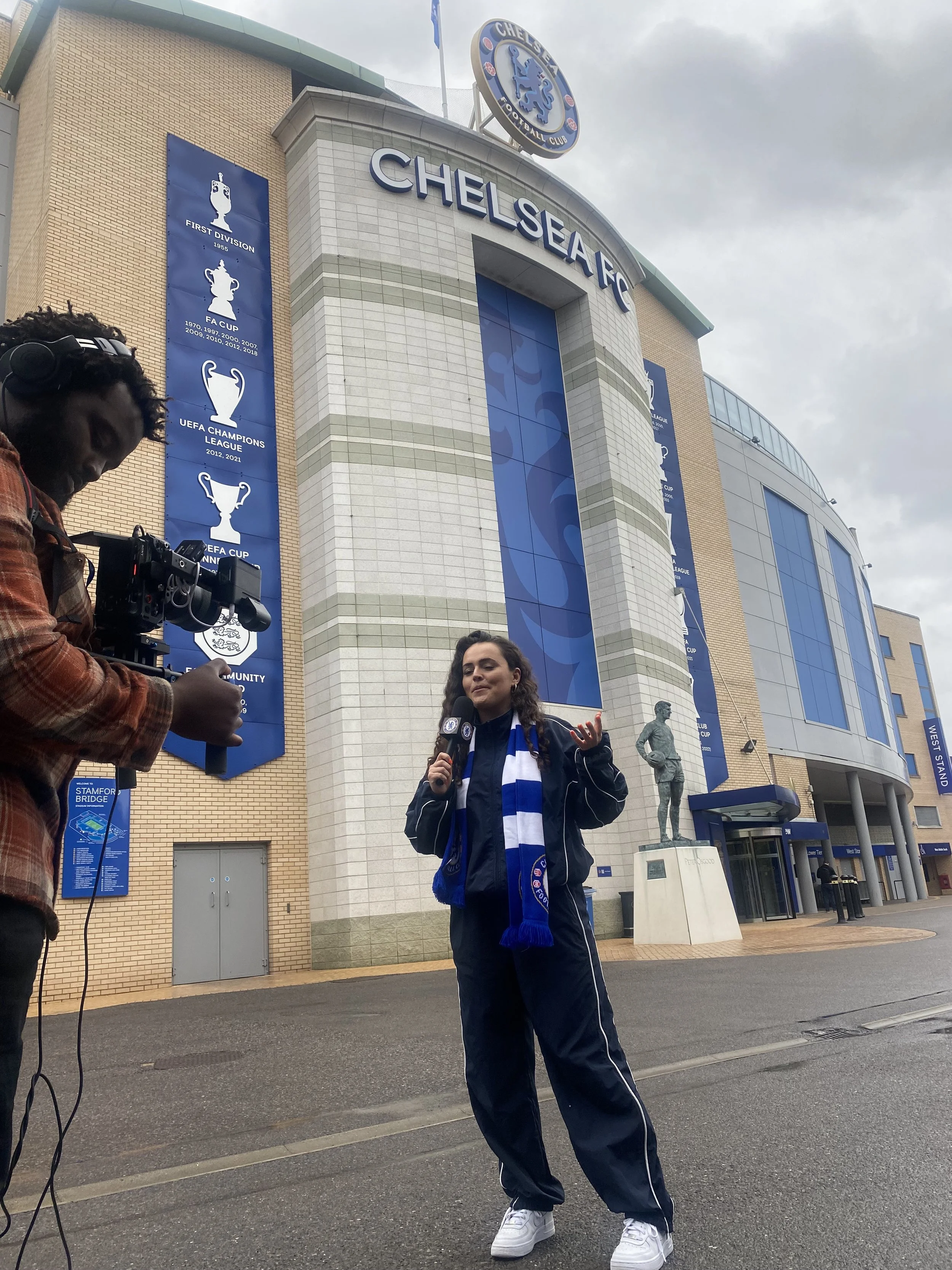 A woman dressed in sportswear holding a microphone stands outside Chelsea Football Club stadium, with a man filming her. The stadium has large blue banners with trophy icons and text listing football achievements, and a statue of a football player is