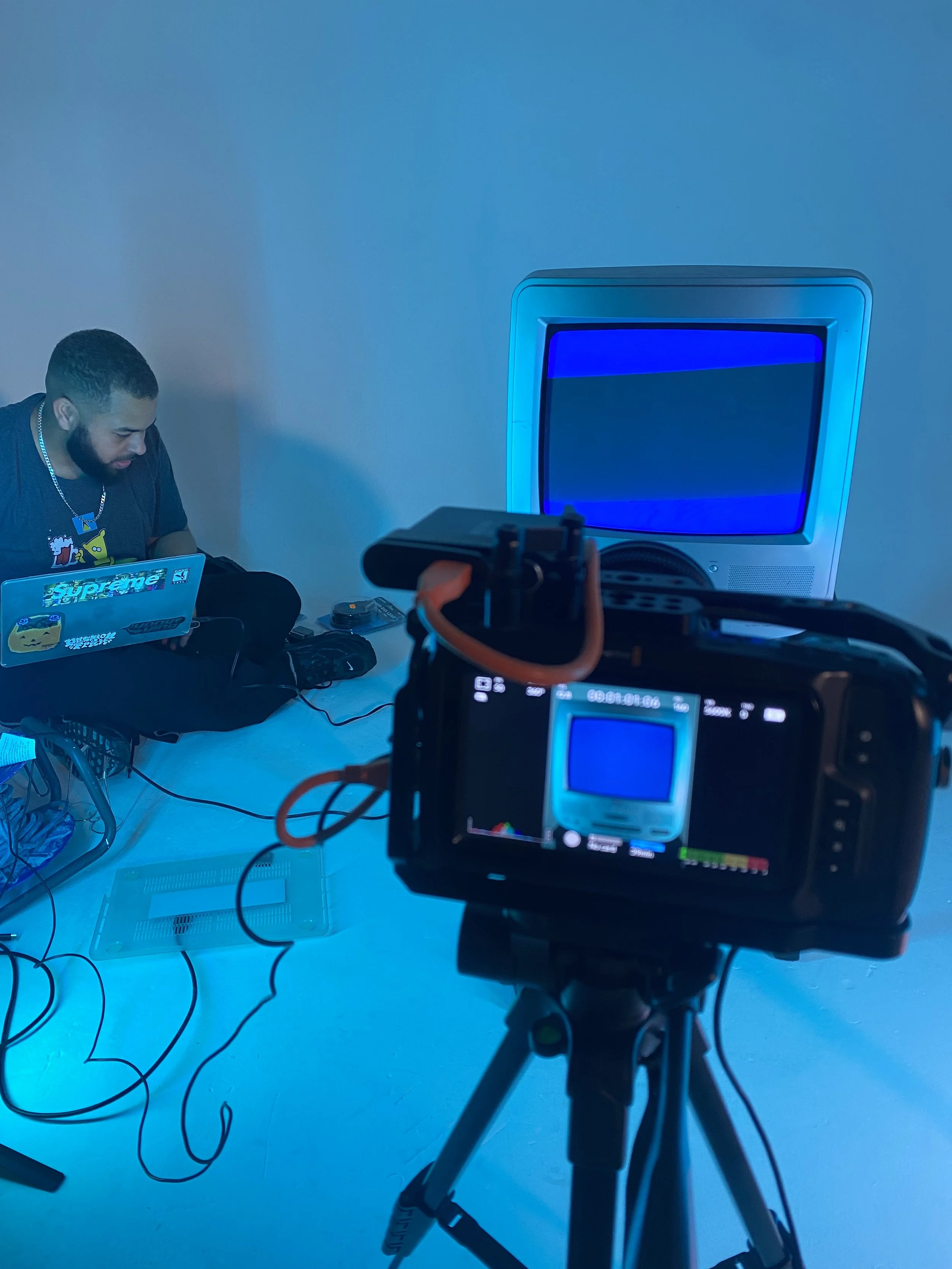 A man sitting on the floor with a laptop, surrounded by cables, in front of an old-fashioned television with a blue screen, recording with a camera on a tripod.