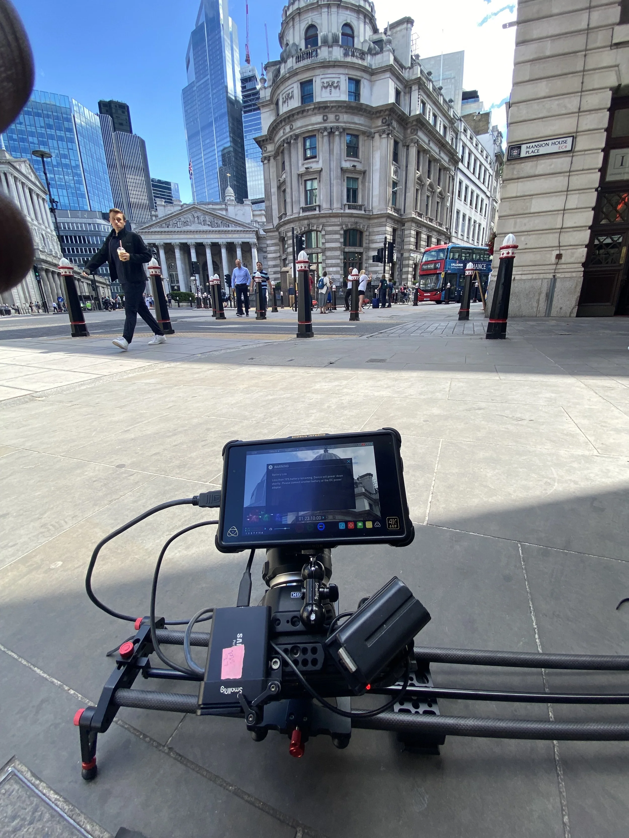 A camera mounted on a rig captures a scene at a busy city street with pedestrians, historic and modern buildings, and a red double-decker bus in the background.