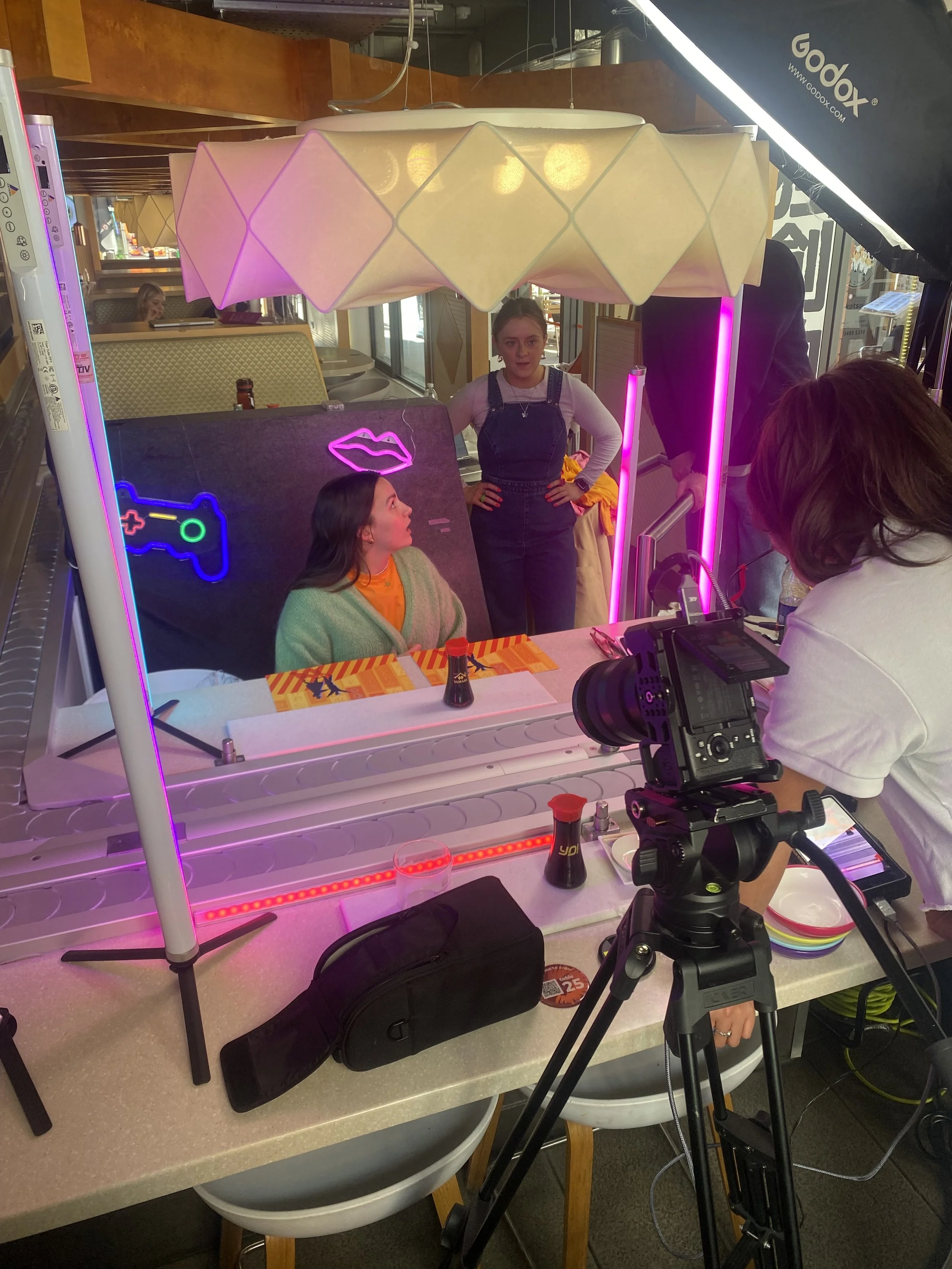 A behind-the-scenes look at a photo shoot or filming of two women in a restaurant. One woman is seated in front of a mirror with neon light outlines of a controller and lips behind her. The other woman stands with hands on hips. A professional camera