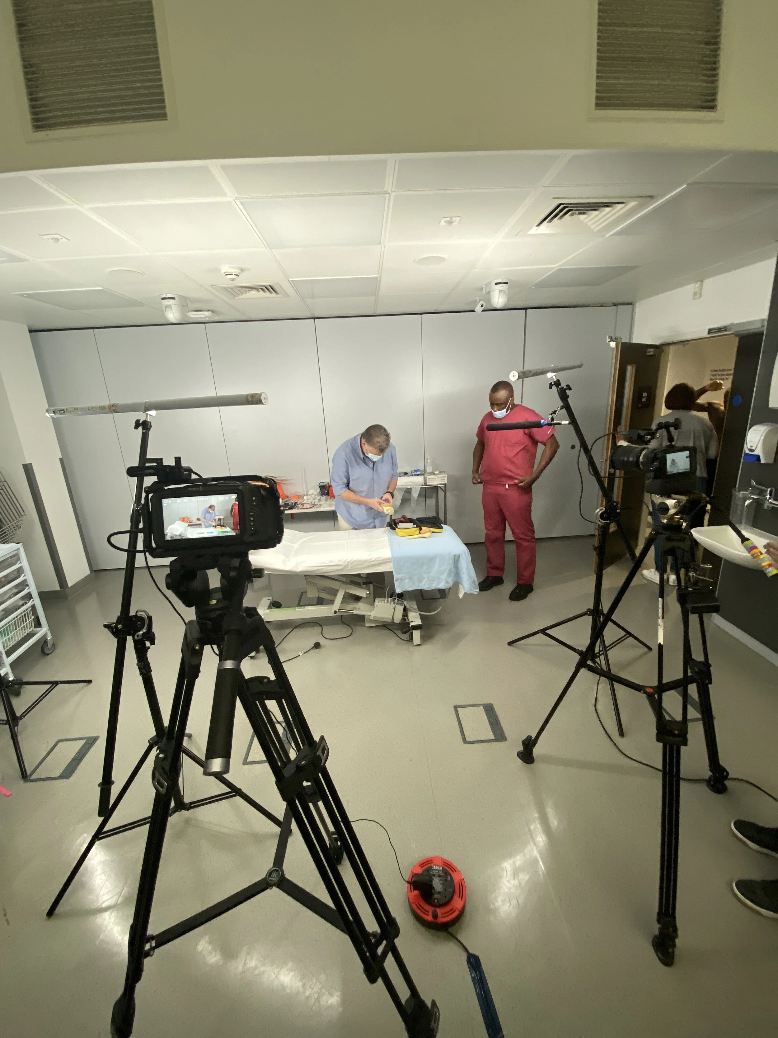 Medical professionals demonstrating medical procedures on a hospital bed, with camera and lighting equipment set up for filming or photography.