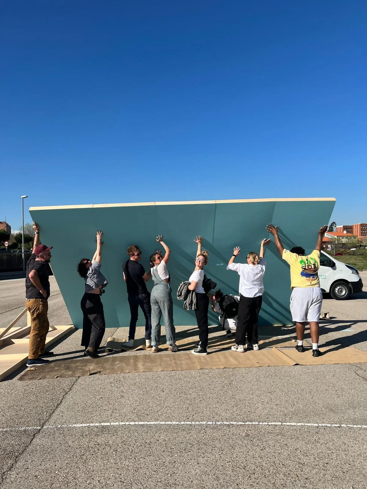 Group of nine people standing in front of a large sculptural wall, holding it up with their hands.