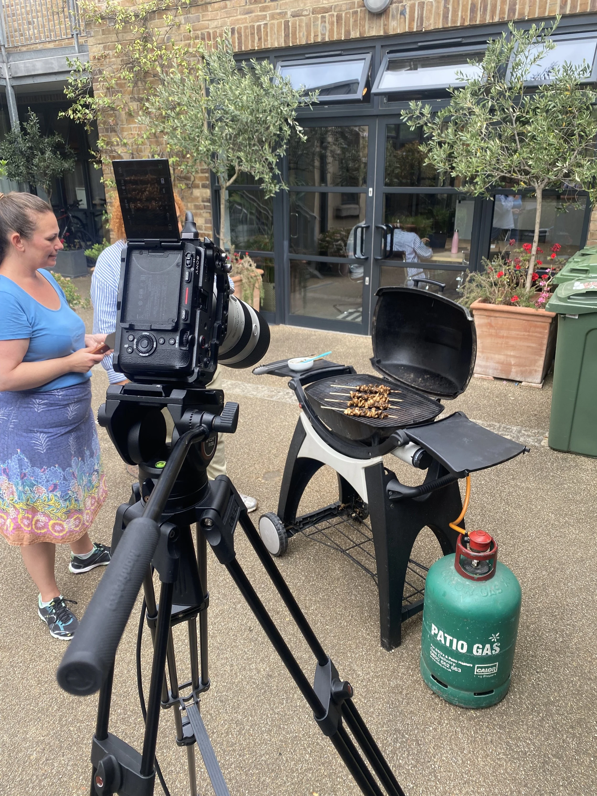 A camera on a tripod is filming a group of people around a barbecue grill with skewers of food outside a building with large glass doors. Two women are visible, one in a blue shirt and another in a striped shirt, standing and talking.