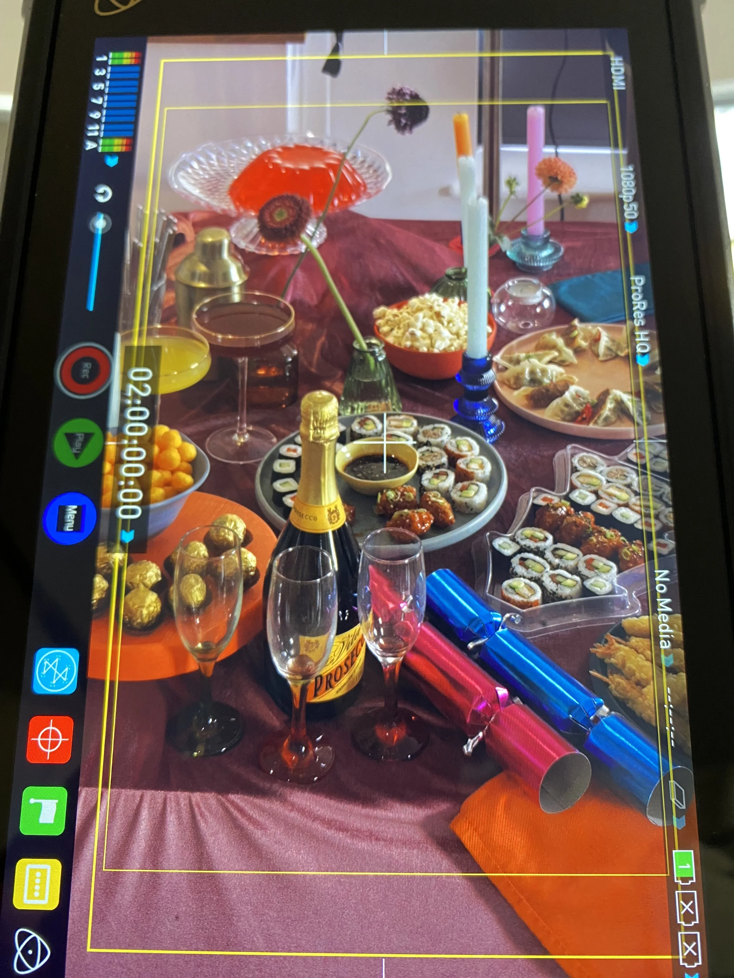 A colorful table with various foods and decorations, including sushi, popcorn, chocolate truffles, and a bottle of Prosecco, arranged with candles, flowers, and party poppers.