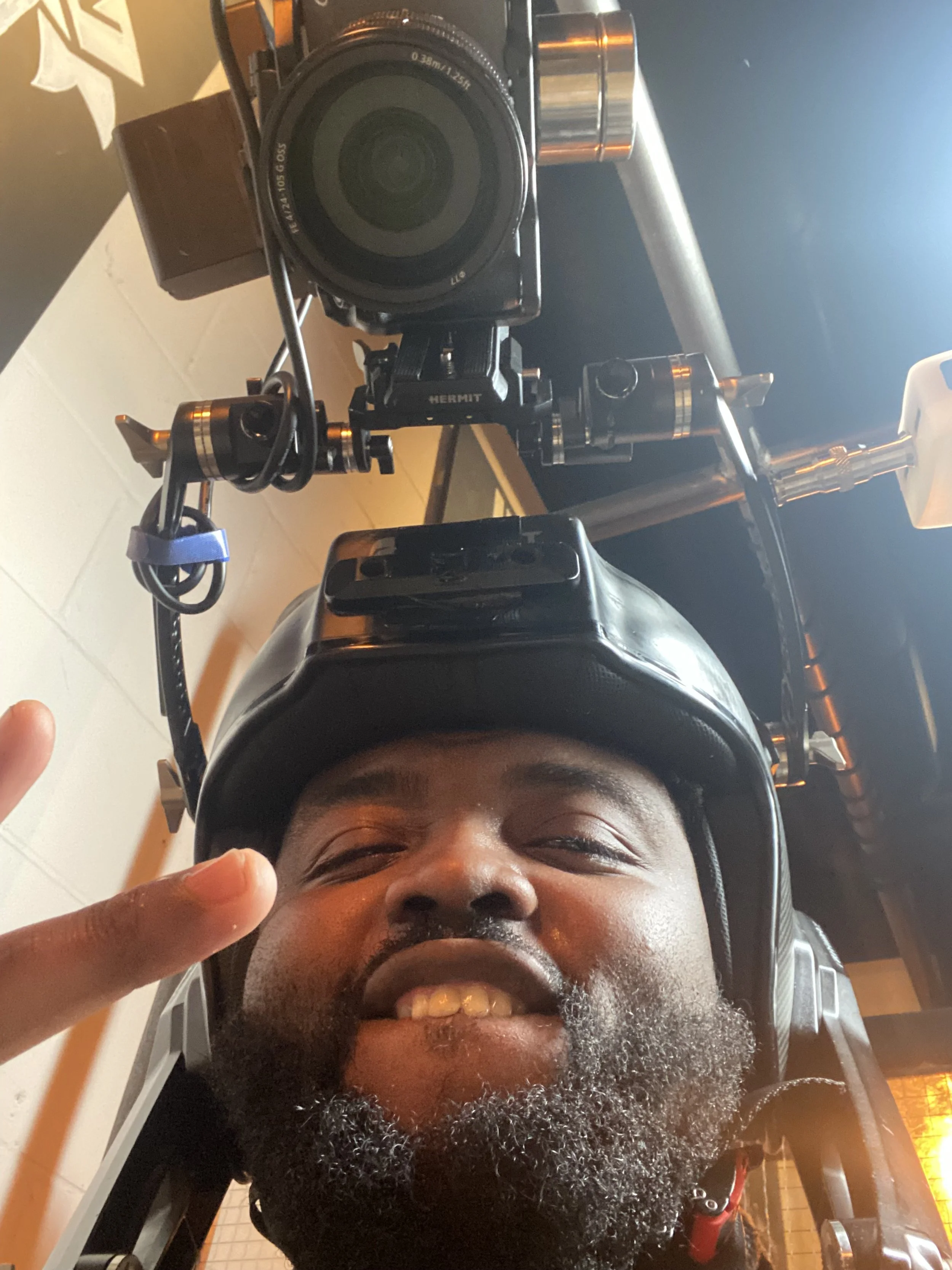 A man wearing a helmet, smiling, taking a selfie from below with a large camera mounted on a stabilizer above him.