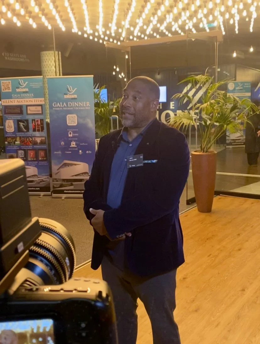 A man in a suit jacket being interviewed at an indoor event with illuminated ceiling lights, banners advertising a gala dinner, and potted plants in the background.