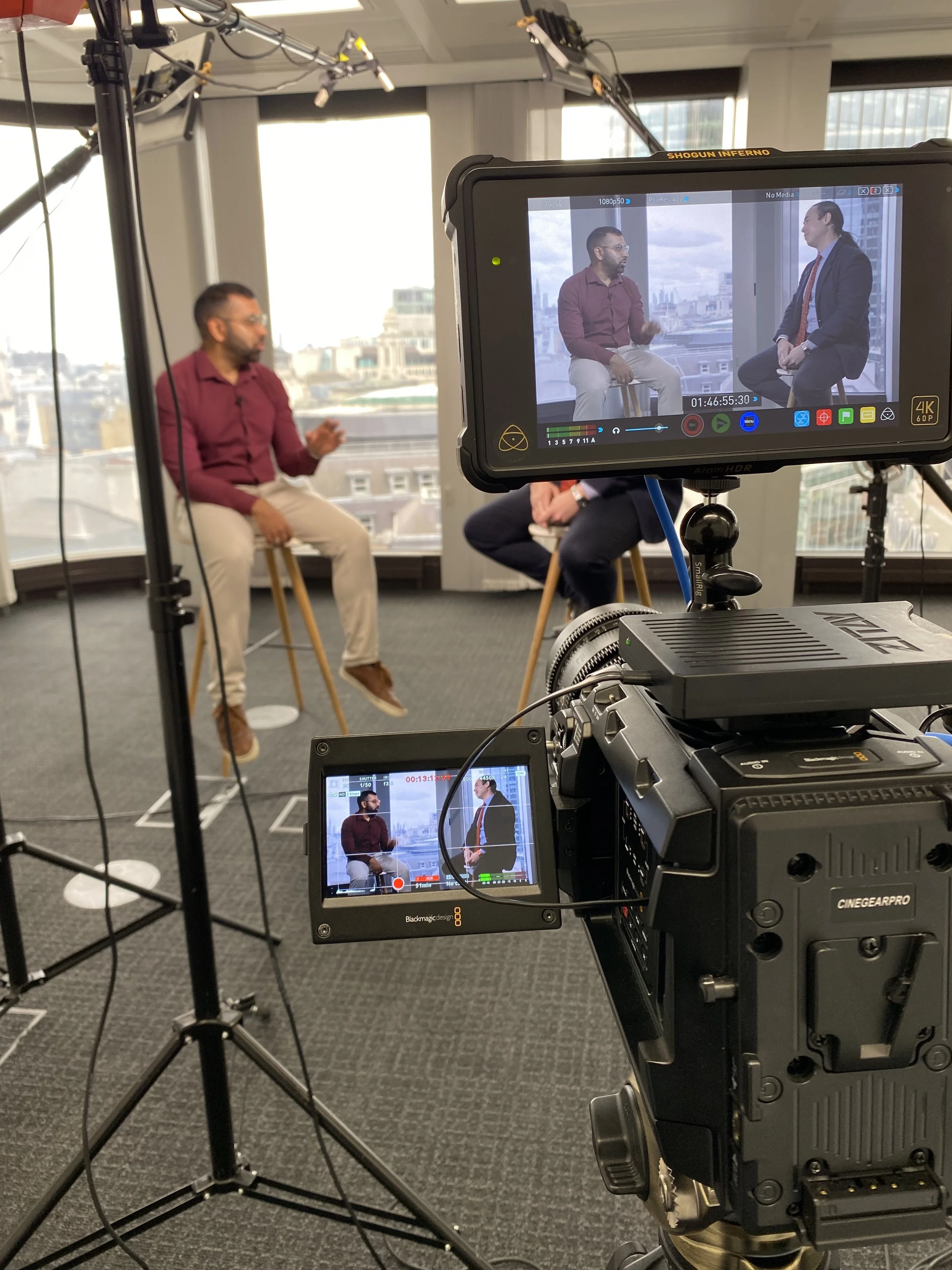 Two men are sitting on stools in an office, engaged in a video interview or discussion, with a city skyline visible through the windows behind them. A professional camera setup is recording them, with two monitors showing their live feed.