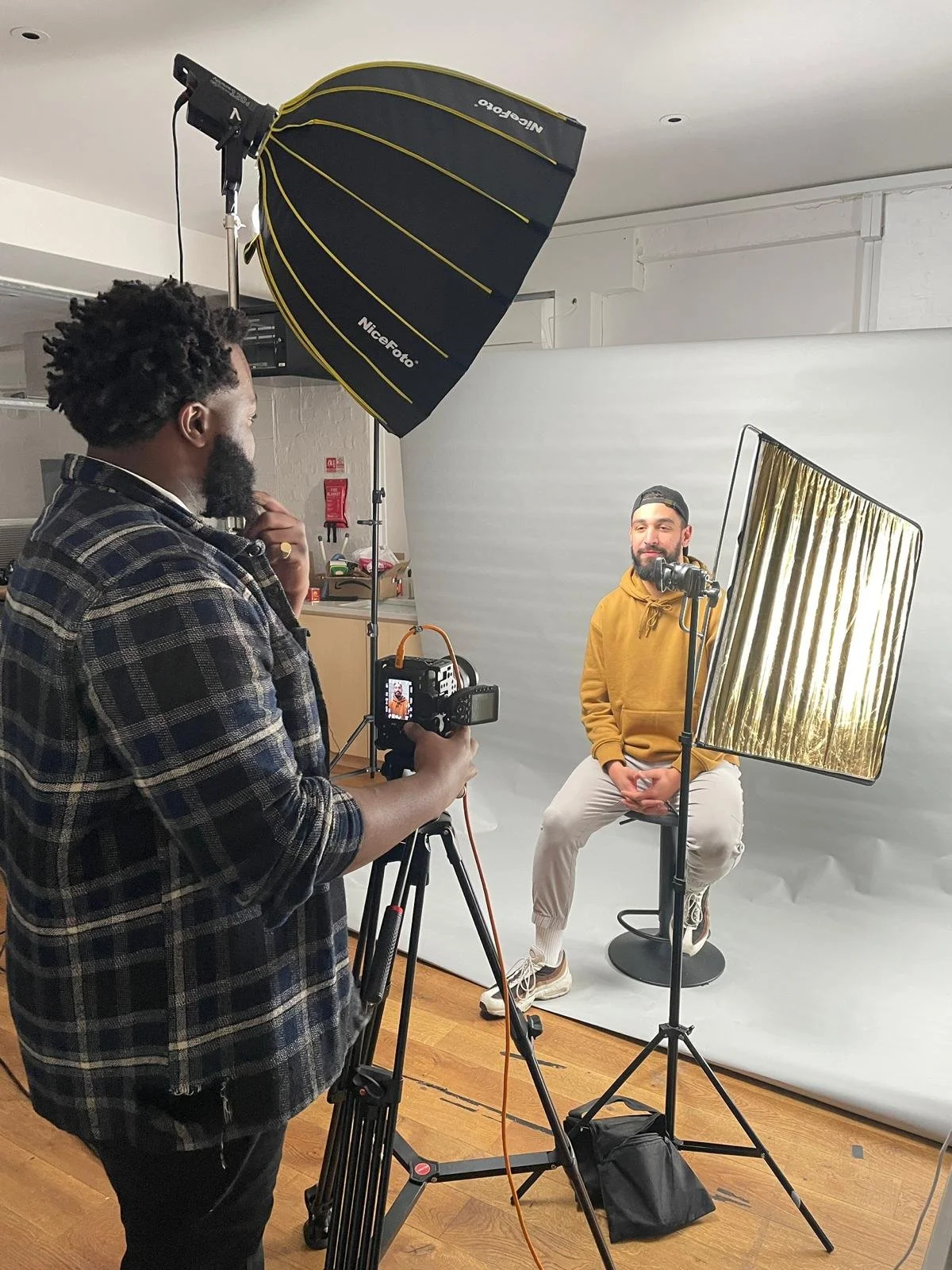 A man in a yellow hoodie sitting on a stool in front of a camera with a studio backdrop and lighting equipment, during a photo or video shoot.
