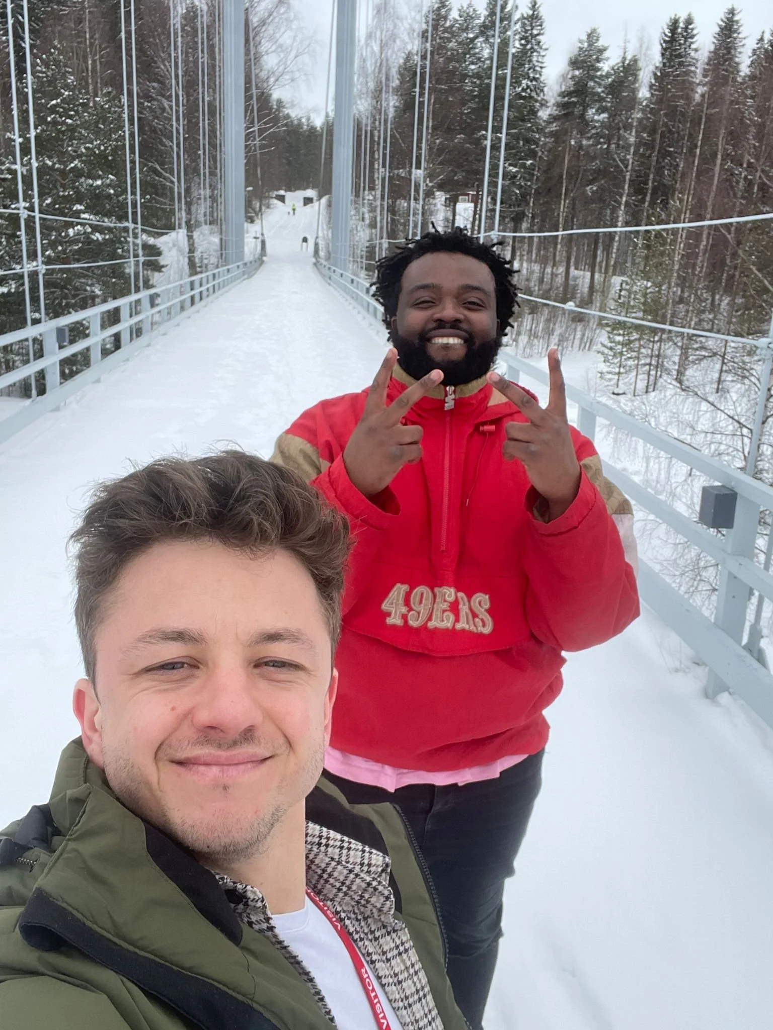 Two men smiling on a snowy suspension bridge with snow-covered trees in the background.