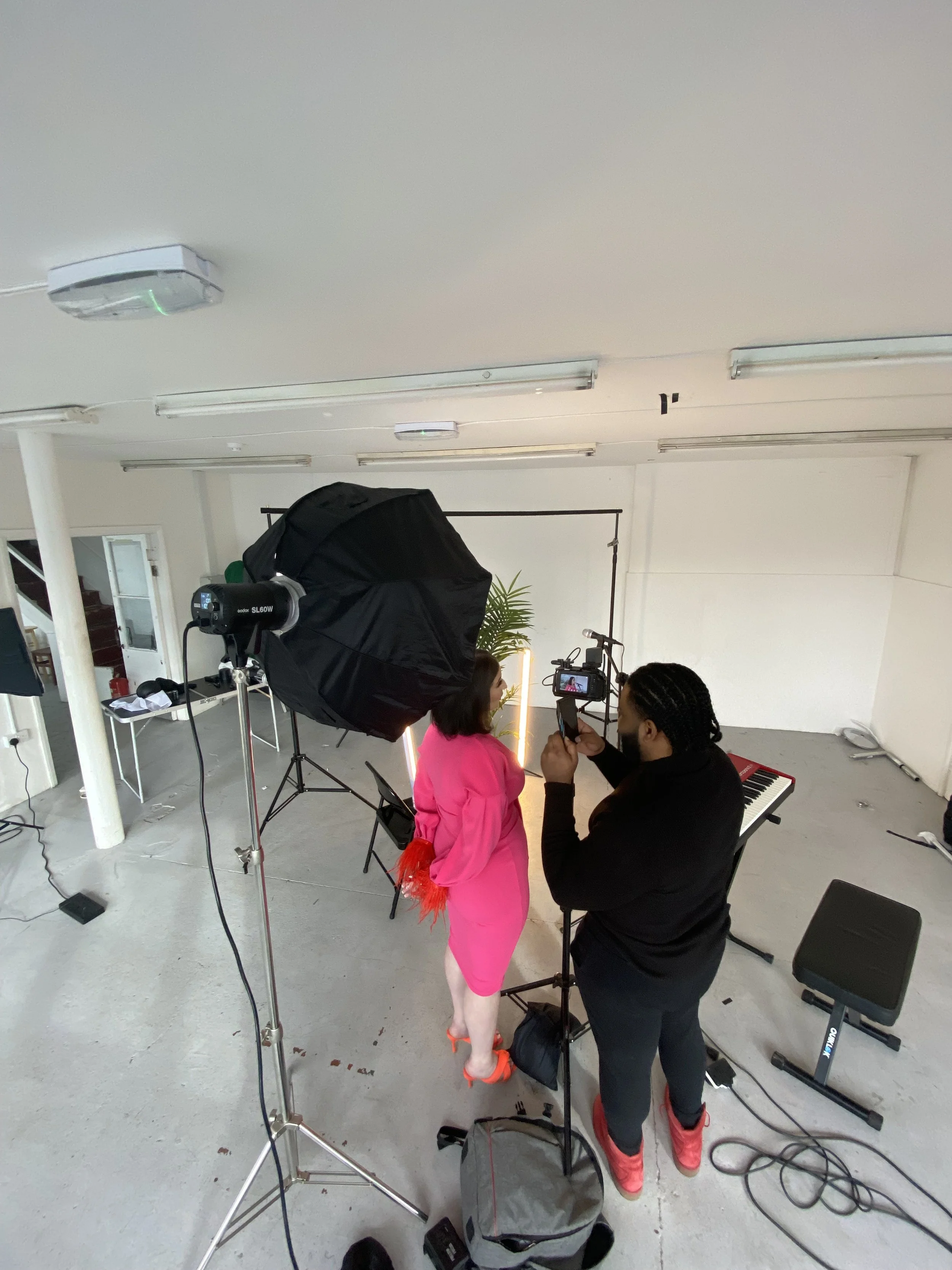 A woman in a pink dress is being photographed or filmed in a studio with lighting equipment and a keyboard in the background.