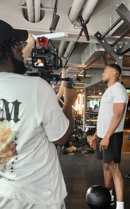 A man in a white t-shirt and black shorts standing in a gym, being filmed or photographed by a person with a camera mounted on a stabilizer. The scene takes place in a modern gym with workout equipment visible in the background.