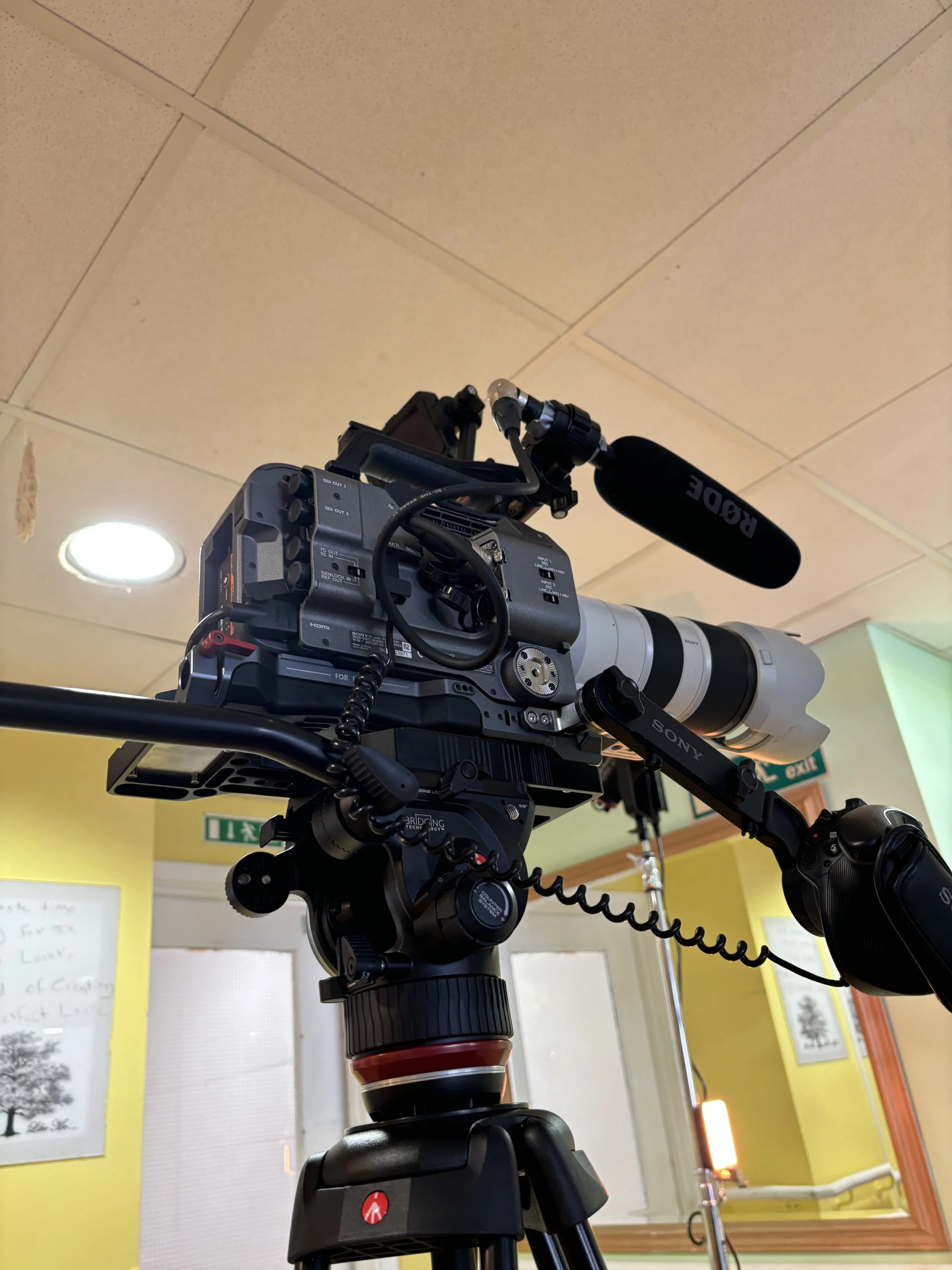 Professional video camera with a large telephoto lens mounted on a tripod in an indoor setting.