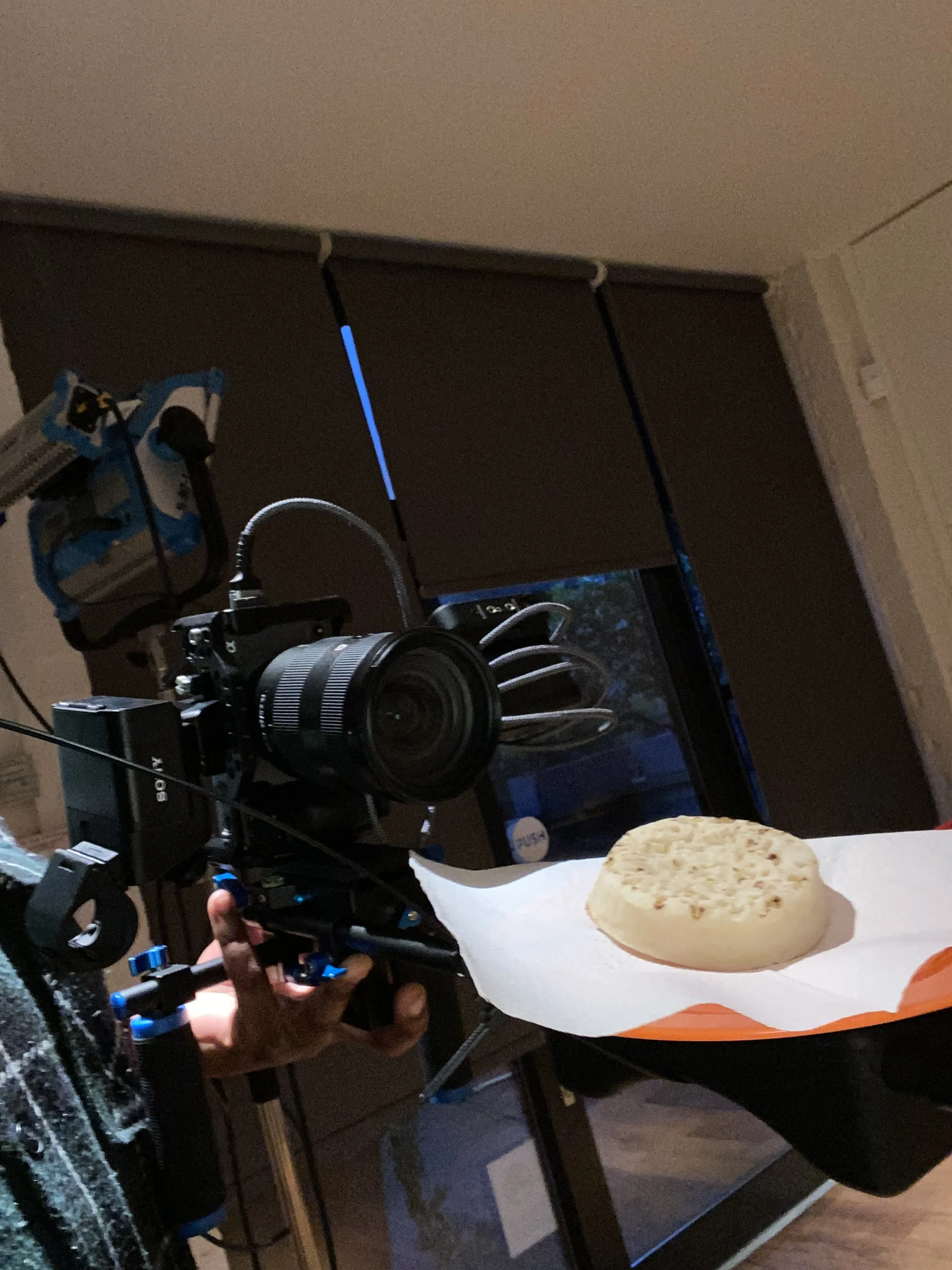 A person is filming food with a professional camera setup indoors. The food appears to be a round, flat, textured snack on a paper-lined tray.